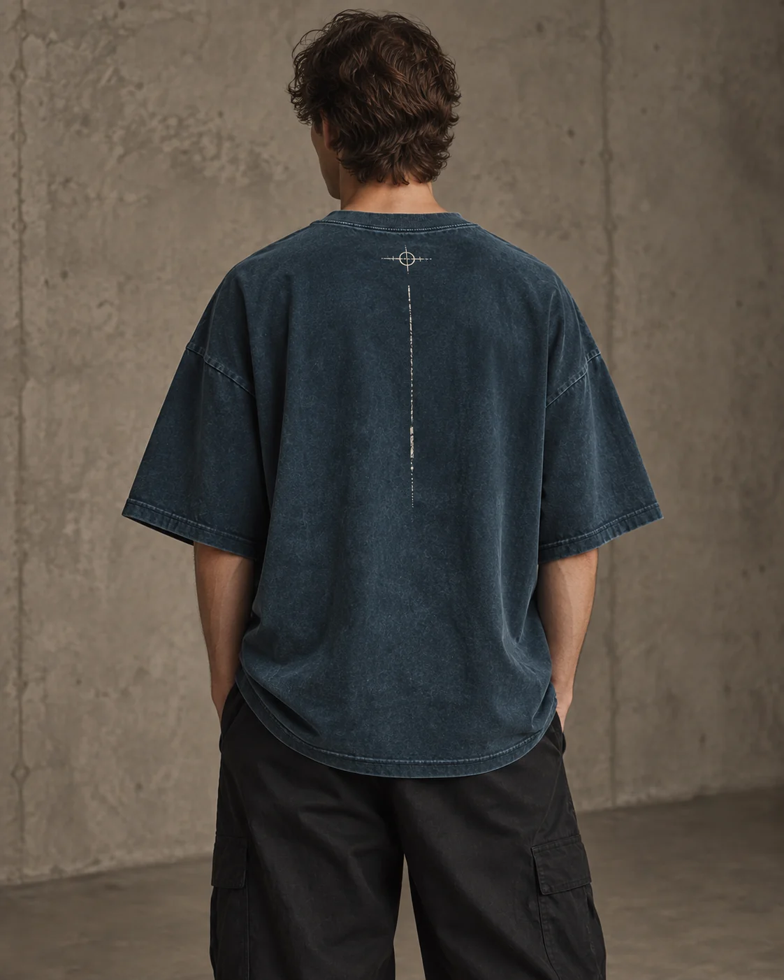 Back view of the same model in petrol-blue oversized T-shirt, with small registration motif and a narrow broken vertical print line